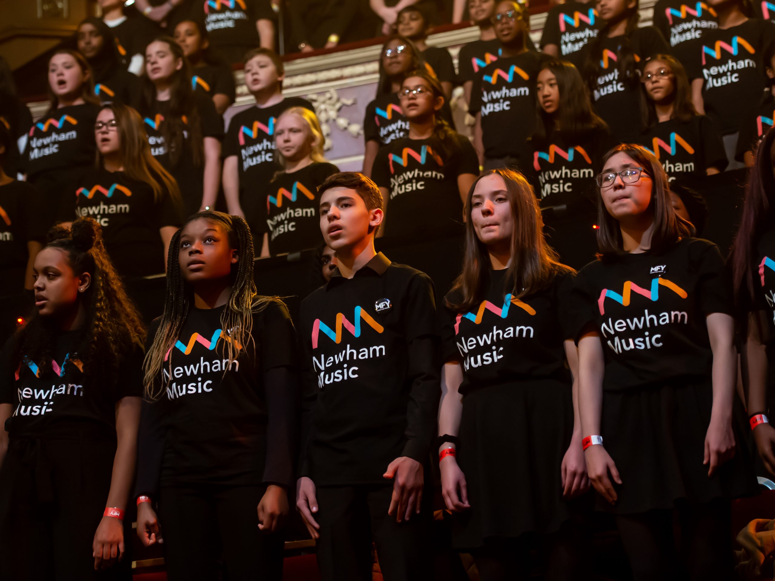 a large group of young people singing wearing black tshirts with Newham Music logo