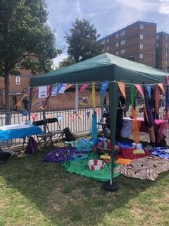 A green gazebo set up on grass with a block of flats in the background. The gazebo is decorated with bunting and there are brightly coloured materials on the ground.