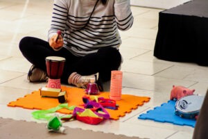 A lady sitting on the floor with a mat in front of her. On the mat are small instruments such as a drum and some sensory toys